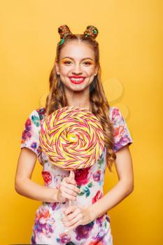 Funny teenage girl holdss big lollipop. Happy teenager with colorful caramel candy, yellow background in studio. Happy smiling girl with bright makeup.