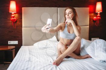 Pretty woman in underwear sitting in bed and makes selfie on phone. Good morning in bedroom