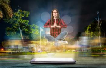 Young woman in yoga pose hovers above a big phone with a glowing screen, evening street with long exposure light effect on background. Communication god