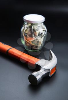 Money saving control concept, hammer against glass jar full of dollars. Cash economy, home bank