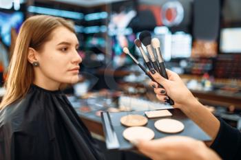 Visagiste hand with brushes against customers face, woman in make-up shop. Female client in beauty salon, makeup procedure