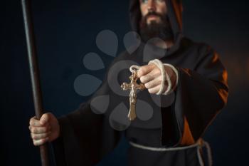 Medieval monk with wooden stick and cross in hands, religion. Mysterious friar in dark cape, Mystery and spirituality