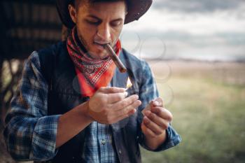 Bearded cowboy lights a cigar with matches, texas ranch on background, western. Vintage male person in leather clothes on farm, wild west culture