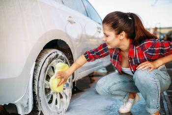 Cute woman with sponge scrubbing vehicle wheel with foam, car wash. Lady on self-service automobile washing. Outdoor carwash at summer day 