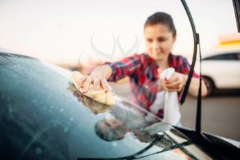 Cute woman cleans front glass of the car with sponge and spray, carwash. Lady on self-service automobile washing. Outdoor vehicle cleaning at summer day 