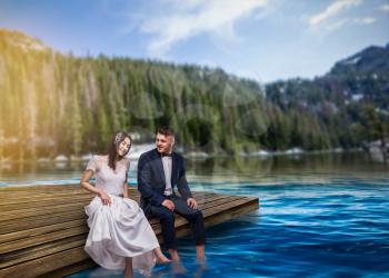 Bride and groom sit on the pier and wet their feet in the water, romantic scene on the lake, mountains and forest on background, summer nature landscape. Honeymoon or wedding trip
