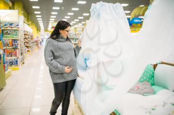 Pregnant woman choosing baby cot in store. Future mother in shop of goods for newburns