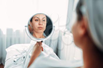 Female patient looks at her face through the mirror, cosmetology clinic. Result of the rejuvenation procedure. Facial skincare in spa salon, beauty medicine