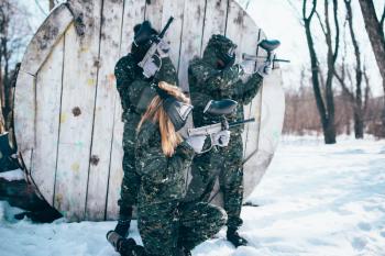 Paintball team in uniform and masks shooting at the enemy, side view, winter forest battle. Extreme sport game