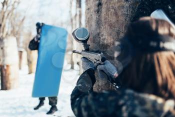 Female paintball player shooting at the enemy with the shield, back view, winter forest battle. Extreme sport game, players fights in protection masks and uniform