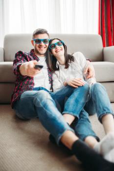 Laughing couple in 3D glasses sitting on the floor against couch and watch TV at home, window on background