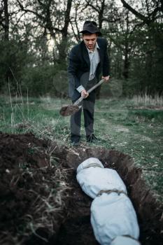 Maniac with a shovel buries victim into a grave, the body wrapped in a canvas, serial murderer concept