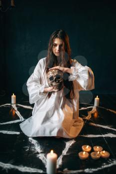Young woman in white shirt holds human skull in hand, pentagram circle with candles, smoke all around, witch