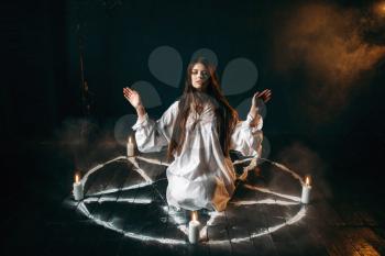 Witch in white shirt sitting in the center of pentagram circle with candles, occult ritual, evocation of spirits. Occultism and exorcism