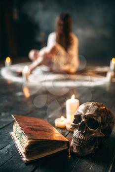 Human skull and old spellbook on black wooden floor, female person in white shirt sitting in pentagram circle with candles on background. Dark magic, occultism and exorcism
