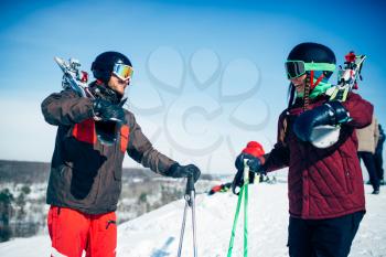 Male and female skiers poses with skis and poles in hands, blue sky and snowy mountains on background. Winter active sport, extreme lifestyle. Downhill skiing