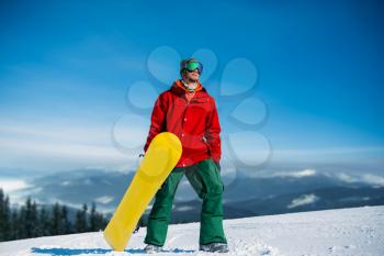 Snowboarder in glasses poses with board in hands, blue sky and snowy mountains on background. Winter active sport, extreme lifestyle, snowboarding