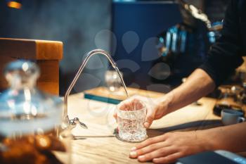 Male barista pours filtered water into the glass, cafe or bar counter on background. Professional coffee preparation by bartender in cafeteria, barman occupation