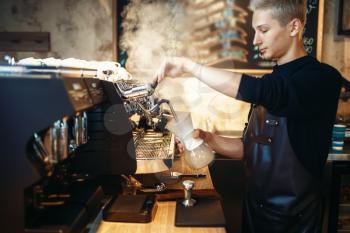 Young male barista in black apron prepares cappuccino on coffee machine. Professional bartender occupation