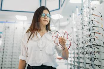 Young female buyer holds many glasses in hand, optics store, showcase with spectacles on background. Professional eyecare in glasses shop concept, eyeglasses choice