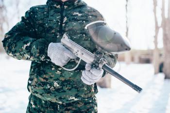 Male paintball player in protection mask and uniform holds marker gun in hands, soldier before winter forest battle. Extreme sport, military game equipment
