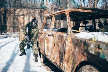 Paintball battle around burned car in winter forest, paintballing. Extreme sport, military game
