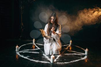 Witch in white shirt holds knife and reads spell, pentagram circle with candles, dark magic ritual process. Occultism and exorcism