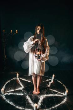 Young woman in white shirt holds kerosene lamp in hand, pentagram circle with candles. Dark magic ritual, occult and exorcism