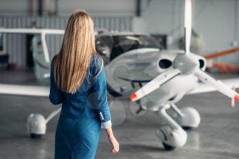 Attractive stewardess in uniform poses against turboprop airplane in hangar. Air hostess in suit near plane. Private airline, flight attendant