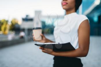Female business person with cardboard coffee cup and notepad outdoors, office building on background. Black businesswoman in skirt and white blouse