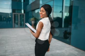 Business woman with notepad outdoors, back view, office building on background. Black businesswoman in skirt and white blouse