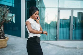 Black business lady talking by mobile phone and looks at the time on the clock in front of office building. Smiling businesswoman in skirt and white blouse, negotiation outdoors