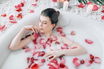 Attractive lady lying in the bath with foam and decorated with rose petals. Full relaxation in bathroom