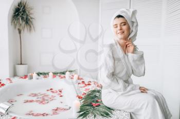 Attractive woman in white bathrobe and towel on head sitting on the edge of the bath decorated with rose petals. Luxury bathroom interior with window on background
