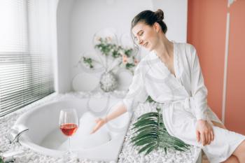 Young woman in white bathrobe sitting on the edge of the bath with foam. Bathroom interior with window and glass with red wine on background