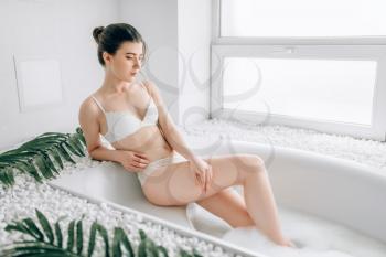 Attractive woman in white underwear sitting in bath with foam. Bathroom interior with window on background