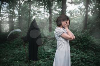 Young female victim in white dress in the forest, death in a black hoodie and with a scythe on background. Photo in horror style, exorcism