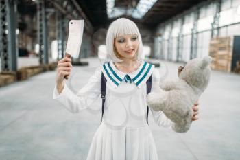 Cute anime style blonde girl with knife and teddy bear in hands. Cosplay woman, japanese culture, doll with toy on abandoned factory