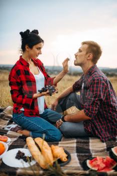 Woman feeds grapes to her man on picnic in summer field. Romantic junket of man and woman, love couple happy together