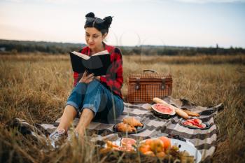 Young woman sitting on plaid and reads book, picnic in summer field. Romantic junket, happy holiday