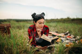 Young woman lies on plaid and reads book, picnic in summer field. Romantic junket, happy holiday