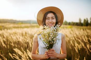 Young woman in white dress and straw hat with a bouquet of wild flowers in a rye field. Cute girl on summer meadow
