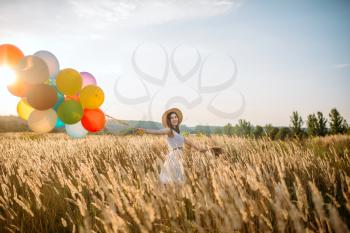 Girl with colorful air balloons walking in wheat field. Pretty woman on summer meadow at sunny day