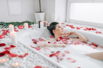 Attractive lady lying in the bath with foam and decorated with rose petals. Full relaxation in bathroom