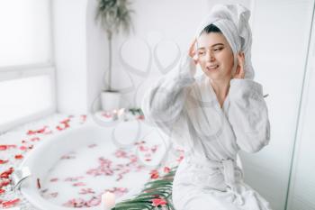 Attractive woman in white bathrobe and towel on head sitting on the edge of the bath decorated with rose petals. Luxury bathroom interior with window on background
