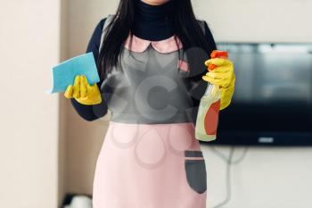 Female cleaner in uniform and gloves holds cleaning equipment in hands, hotel room interior. Professional housekeeping, charwoman