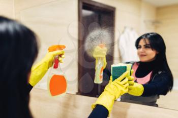 Housemaid hands in rubber gloves cleans the mirror with a cleaning spray, hotel bathroom interior on background. Professional housekeeping service, charwoman, sanitary processing