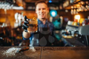 Male bartender in apron holds out a fresh alcoholic beverage at the bar counter. Barman occupation, bartending