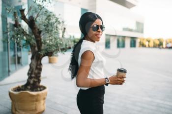 Successful business woman in sunglasses with cardboard coffee cup outdoors, office building on background. Black businesswoman in skirt and white blouse