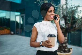 Young business woman with cardboard coffee cup and mobile phone against office building on background. Smiling black businesswoman in skirt and white blouse